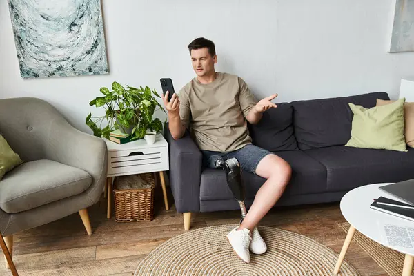 Joven guapo con una pierna protésica hablando en su teléfono inteligente mientras está sentado en el interior. - foto de stock