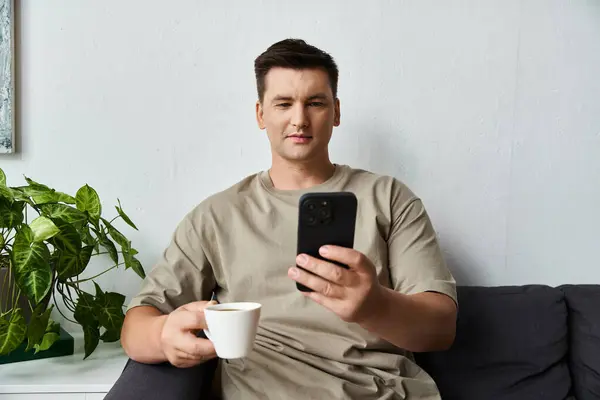 Young man enjoys a warm drink and looks at his phone on a couch. — Stock Photo