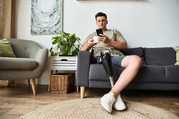 Young man sits comfortably on a couch, enjoying a drink and looking at his phone. — Stock Photo