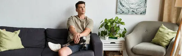 A young man sits on a couch, comfortably dressed, showing his prosthetic leg while relaxing. — Stock Photo