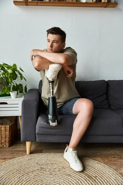 Ein hübscher junger Mann sitzt gemütlich auf einer Couch und umarmt sein Bein mit einer Prothese. — Stockfoto