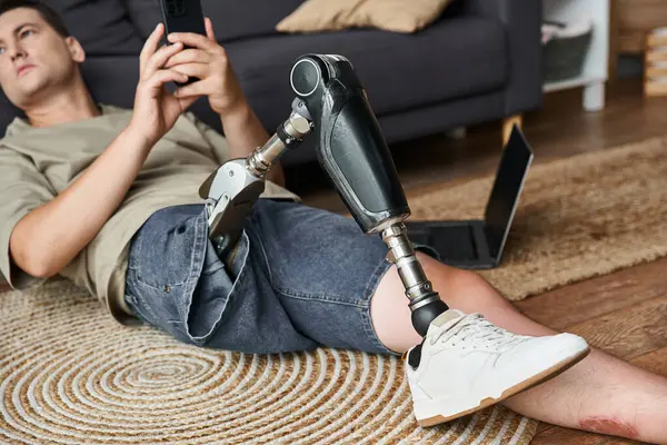 Young man comfortably sitting on a rug, focused on his smartphone, showcasing his prosthetic leg. — Stock Photo