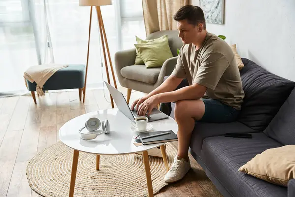Young man sits on couch, using laptop while enjoying a cup of coffee in cozy setting. — Stock Photo