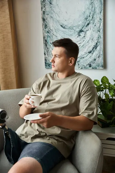 Handsome young man sits on a sofa with a cup of coffee and a plate, looking thoughtful and relaxed. — Stock Photo