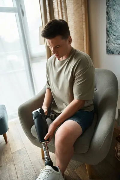 Handsome young man sits in a chair adjusting his prosthetic leg with focus and determination. — Stock Photo
