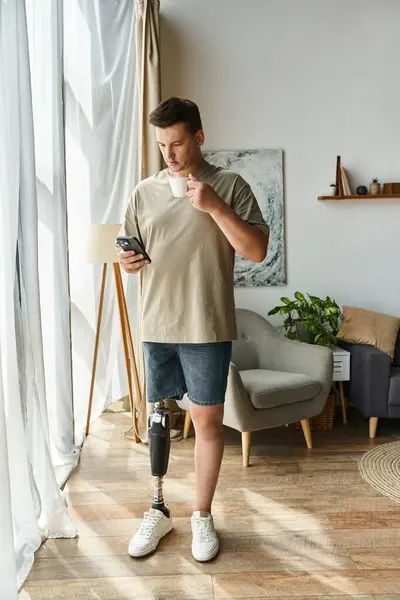 Handsome young man stands by the window checking his phone, showcasing his prosthetic leg. — Stock Photo