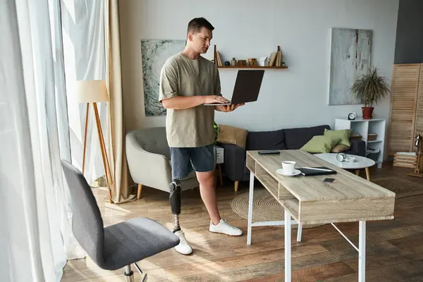 Handsome young man stands at a desk using a laptop in a stylish living room. — Stock Photo