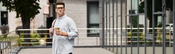 Handsome young man with glasses stands outside holding a coffee cup in a stylish area. — Stock Photo