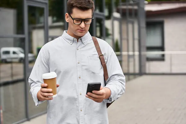Young man with glasses stands outdoors, focused on his smartphone and holding coffee. — Stock Photo
