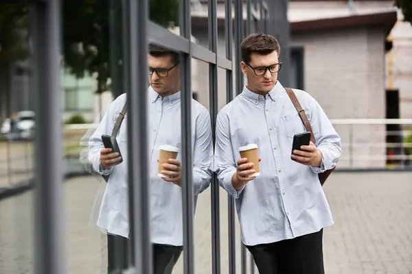 Handsome young man with glasses holds coffee and smartphone, leaning against glass — Stock Photo