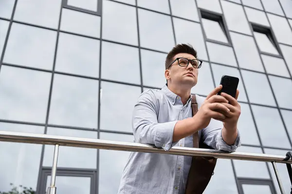 Un joven con gafas interactúa con su smartphone cerca de un edificio moderno. - foto de stock