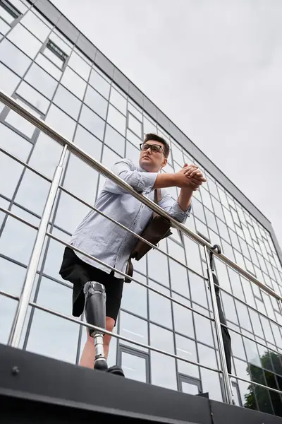 Un giovane uomo con una gamba protesica si erge su un balcone che si affaccia su un paesaggio urbano, perso nel pensiero. — Foto stock