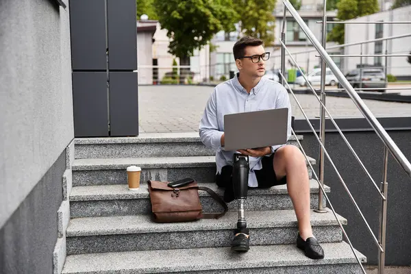 Young man with a prosthetic leg sits on steps, focused on his laptop while enjoying a coffee. — Stock Photo