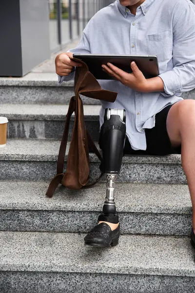 Un joven con una pierna protésica se sienta en escaleras de piedra mientras usa una tableta y sostiene una bolsa. - foto de stock