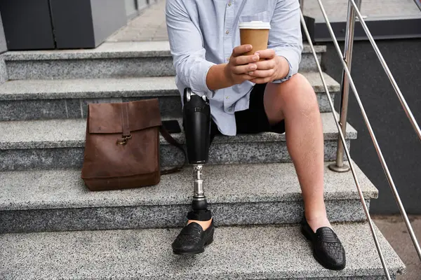 Young man sits on stairs with a coffee cup, showcasing his prosthetic leg and casual style. — Stock Photo