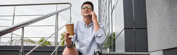 Joven con una pierna protésica se sienta en los escalones, hablando por teléfono y bebiendo café tranquilamente. - foto de stock