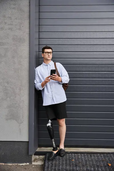A young man with a prosthetic leg uses his smartphone while leaning against a wall. — Stock Photo