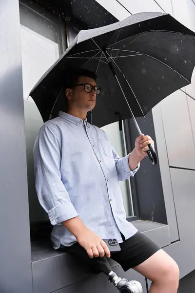 Young man with a prosthetic leg sits by a rainy window, holding an umbrella. — Stock Photo