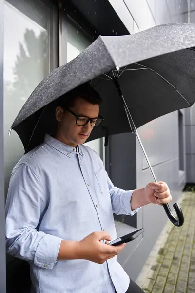 Handsome young man with a glasses uses his phone while sheltered from the rain. — Stock Photo