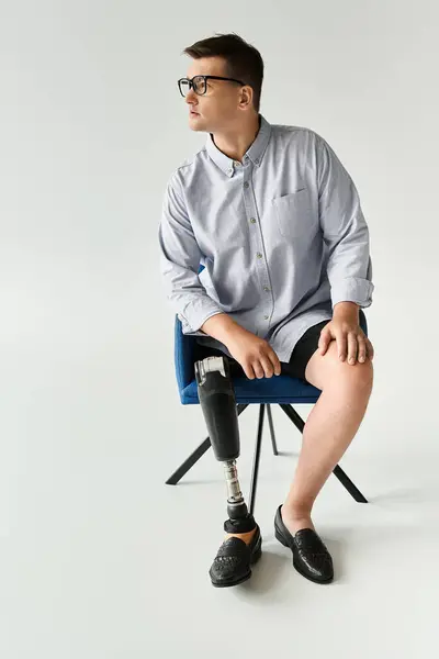 Young man with glasses and a prosthetic leg poses in a stylish chair in a simple studio. — Stock Photo