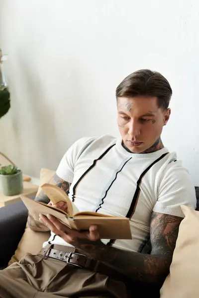 Молодий чоловік з татуюваннями сидить зручно на дивані, занурений у книгу в приміщенні. — Stock Photo