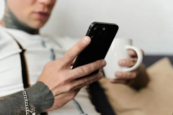 Handsome young man with tattoos relaxes on a couch, sipping coffee and using his smartphone. — Stock Photo