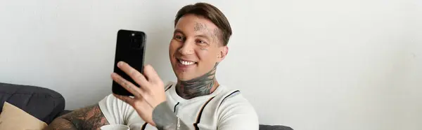 Handsome young man with tattoos smiles while holding his smartphone and sitting comfortably. — Stock Photo