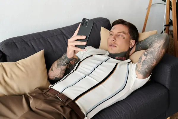 Man with tattoos lounges casually on a couch while checking his smartphone in a cozy room. — Stock Photo