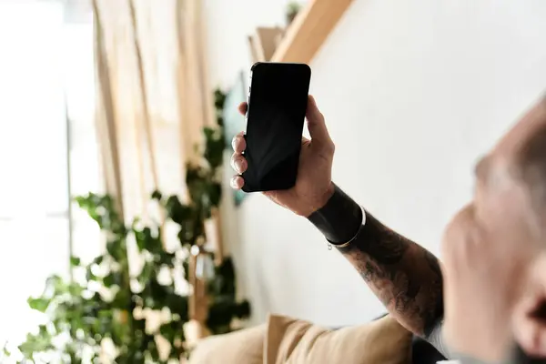 Young man with tattoos enjoys using his smartphone while relaxing indoors with plants. — Stock Photo