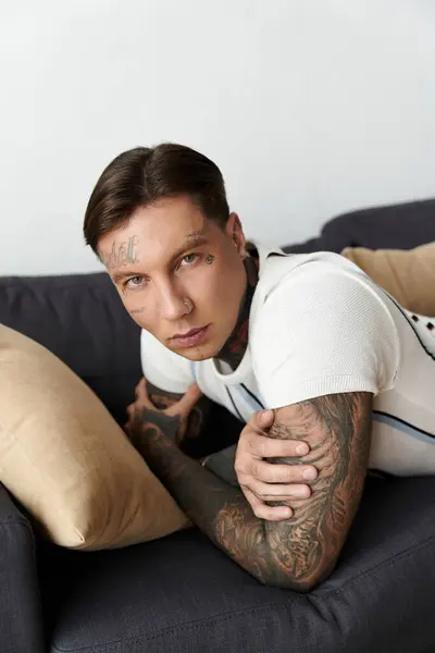 Handsome young man with tattoos lounges comfortably on a couch with cushions. — Stock Photo
