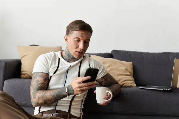 Jeune homme tatoué assis sur le canapé, concentré sur le smartphone et sirotant une tasse dans un cadre décontracté. — Photo de stock