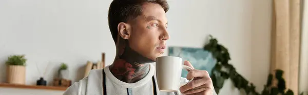 A confident young man with tattoos enjoys a warm cup of coffee while relaxing indoors. — стокове фото