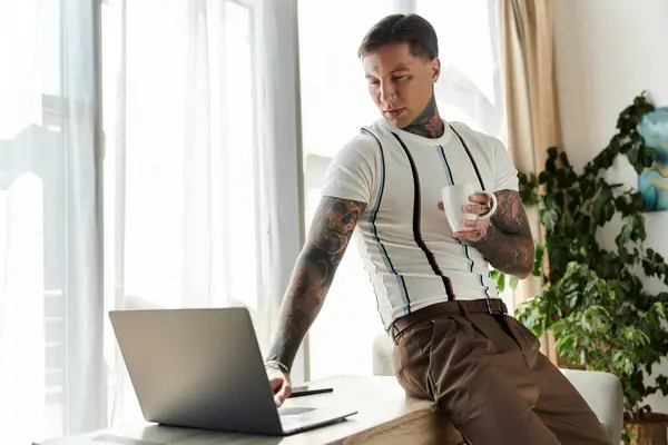 Handsome young man with tattoos sits casually at a desk sipping coffee while focused on laptop. — Stock Photo