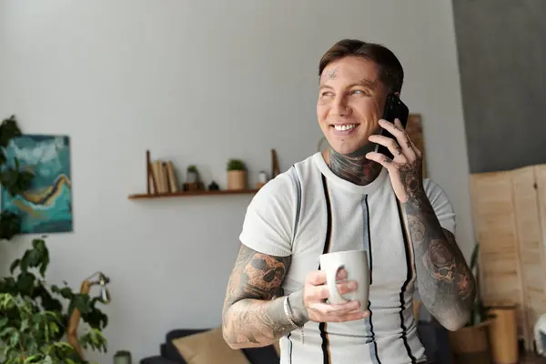 Jovem com tatuagens sorri enquanto fala no telefone e segura uma xícara de café. — Fotografia de Stock