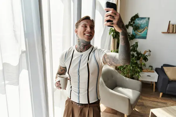 Handsome young man with tattoos smiles while taking a selfie and holding a cup of coffee. — Stock Photo