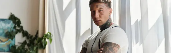 Schöner junger Mann steht mit verschränkten Armen und zeigt Tätowierungen in einem sonnenbeschienenen Raum. — Stockfoto