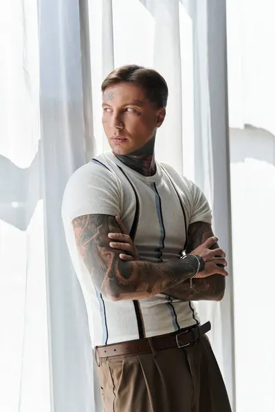 Man stands confidently with arms crossed, showcasing tattoos in a well lit space. — Stock Photo