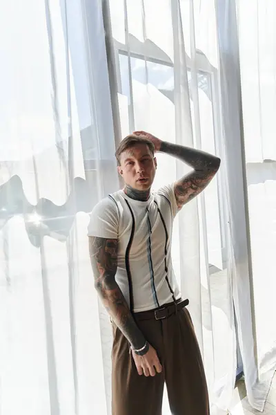 Young man stands confidently by a large window, showcasing his tattoos and fashion sense. — Stock Photo