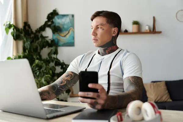Un joven con tatuajes enfocado en su portátil mientras sostiene su teléfono inteligente en una habitación elegante. - foto de stock