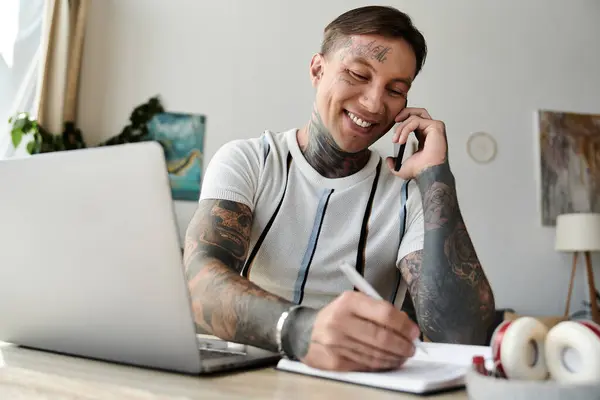Handsome young man with tattoos engages in a phone call while jotting down notes at his desk. — Stock Photo