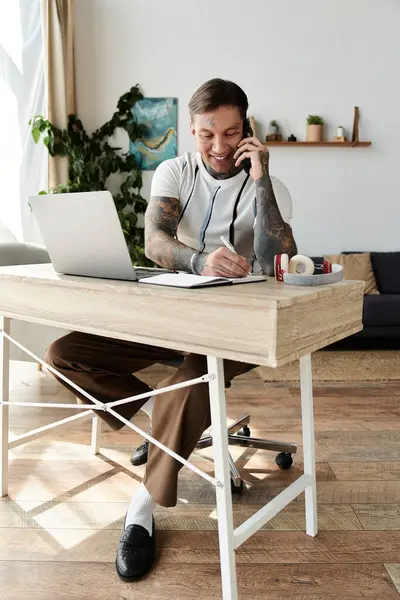 A young man with tattoos smiles while taking a call at his home office desk and writing notes. — Stock Photo