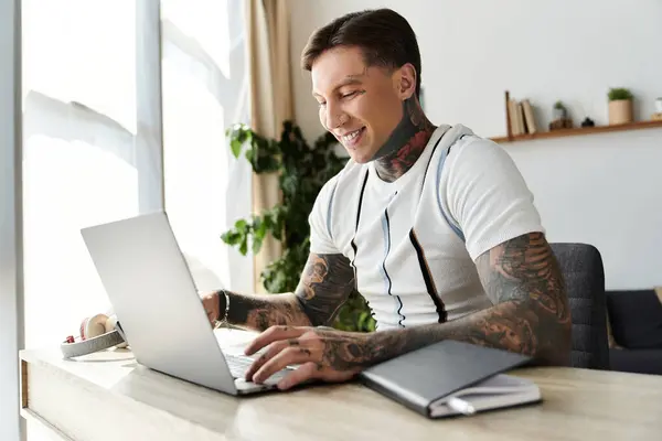 Man with tattoos sits at a desk smiling while typing on a laptop in a stylish setting. — Stock Photo