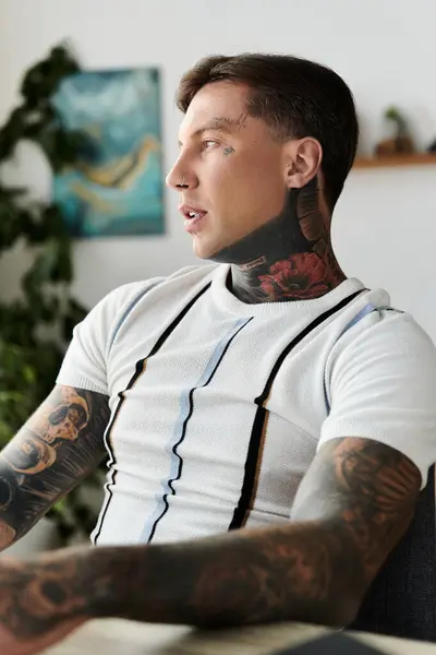 A young man adorned with tattoos is seated, gazing pensively in a contemporary interior. — Stock Photo