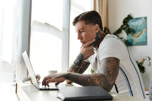 A young handsome man with tattoos is deeply focused while working on his laptop in a bright room. — Stock Photo