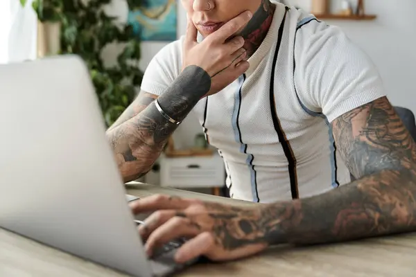 Guapo joven con tatuajes se dedica a un trabajo reflexivo en un ordenador portátil en casa. - foto de stock