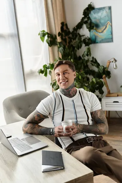 Handsome young man with tattoos sits comfortably at a desk holding a cup of coffee. — Stock Photo