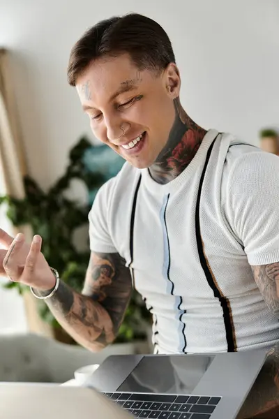 Handsome young man with tattoos smiles while engaged with a laptop in a cozy indoor setting. — Stock Photo