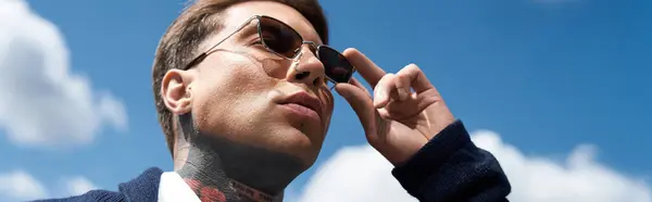 Tattooed man looks up at the sky, adjusting his sunglasses against a bright blue background. — Stock Photo