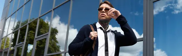 Stylish young man with tattoos adjusts sunglasses near reflective glass building. — Stock Photo