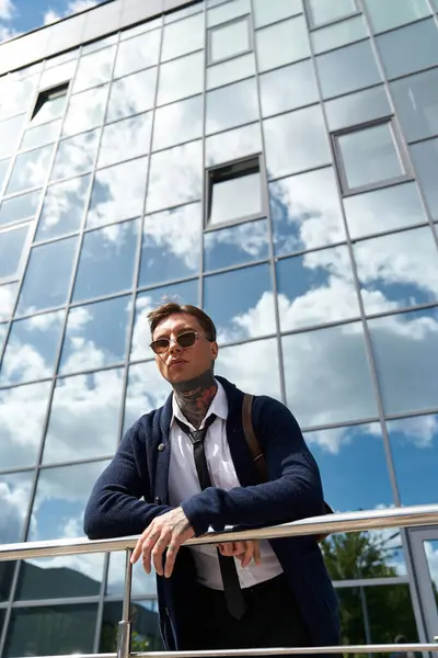 A young man with tattoos stands against a striking glass building reflecting clouds. — Stock Photo